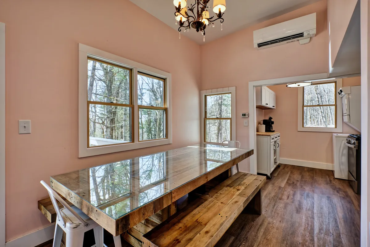 This interior shot captures a spacious dining area featuring a large wooden table beneath a charming chandelier. Multiple windows line the walls, filling the space with natural light and offering beautiful views of the surrounding trees—suggestive of the mountain vistas described. The setup is conducive to family gatherings and laughter-filled meals.