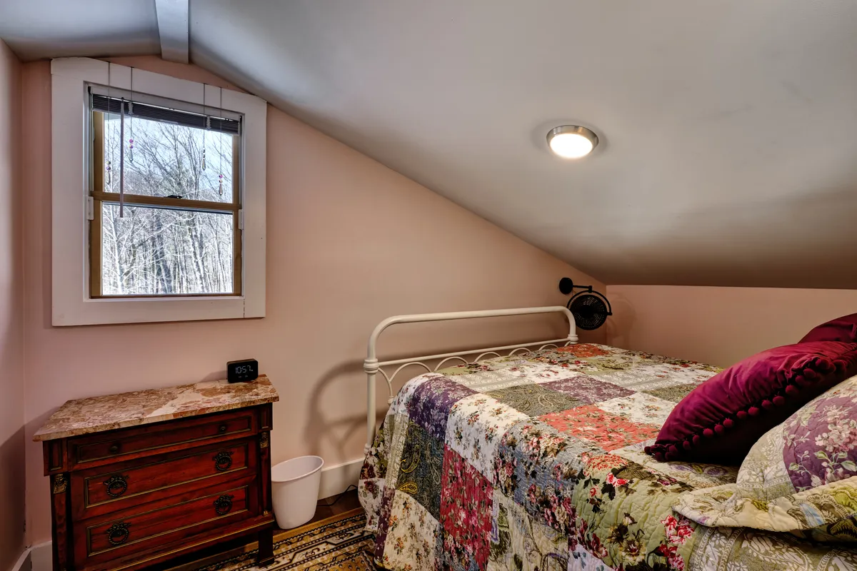 A tranquil bedroom scene featuring a quilted bed with soft, layered linens and a wooden nightstand. Gentle natural light streams in through a window that frames soothing greenery, inviting a sense of peaceful retreat. A book rests on the nightstand, hinting at leisurely moments spent reading or simply soaking in the serene atmosphere.