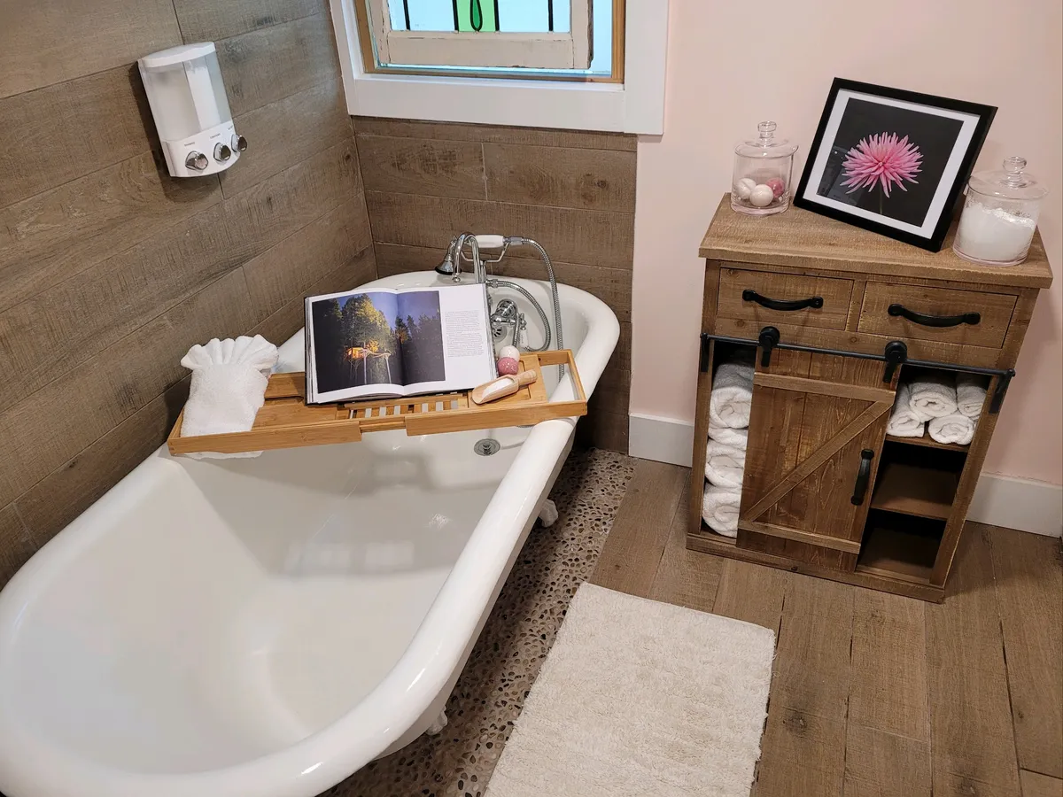 An inviting bathroom with a classic claw foot tub, a wooden tray set with a book, and a decorative cabinet. Sunlight filters through a window with stained glass accents, creating a relaxing, spa-like atmosphere perfect for soaking away stress and embracing mountain serenity.