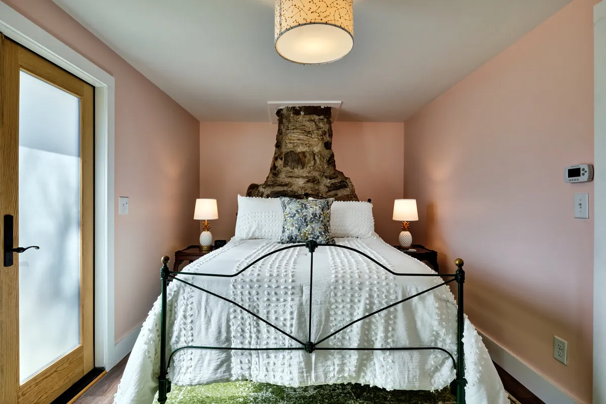 A serene bedroom scene featuring a comfortable bed adorned with decorative pillows, a rustic wooden door, and a natural rock feature incorporated into the room’s design. The ambiance is peaceful and inviting, with soft lighting evoking a sense of relaxation and warmth.