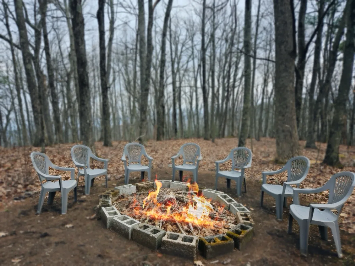 A warmly lit fire pit surrounded by comfortable chairs arranged in a circle, set amidst towering trees and a forest floor scattered with fallen leaves. The setting exudes an inviting, communal atmosphere perfect for evening gatherings, marshmallow toasting, and storytelling under the stars.