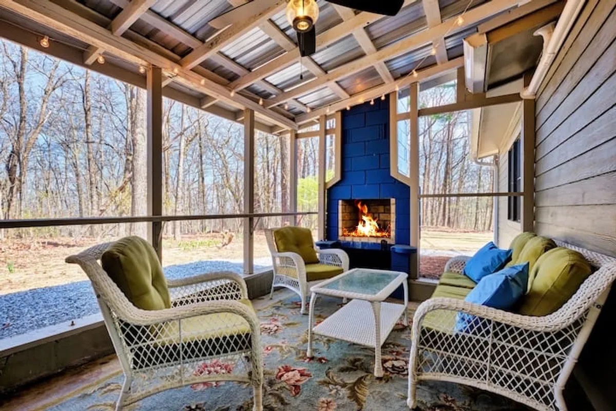 The image showcases a welcoming interior living space centered around a striking stone fireplace. The seating area is adorned with a decorative rug and comfortable chairs, all bathed in warm, natural light. Large windows offer glimpses of the outdoors, enhancing the sense of connection to the surrounding landscape. The mood is cozy and communal, ideal for both relaxation and social gatherings.